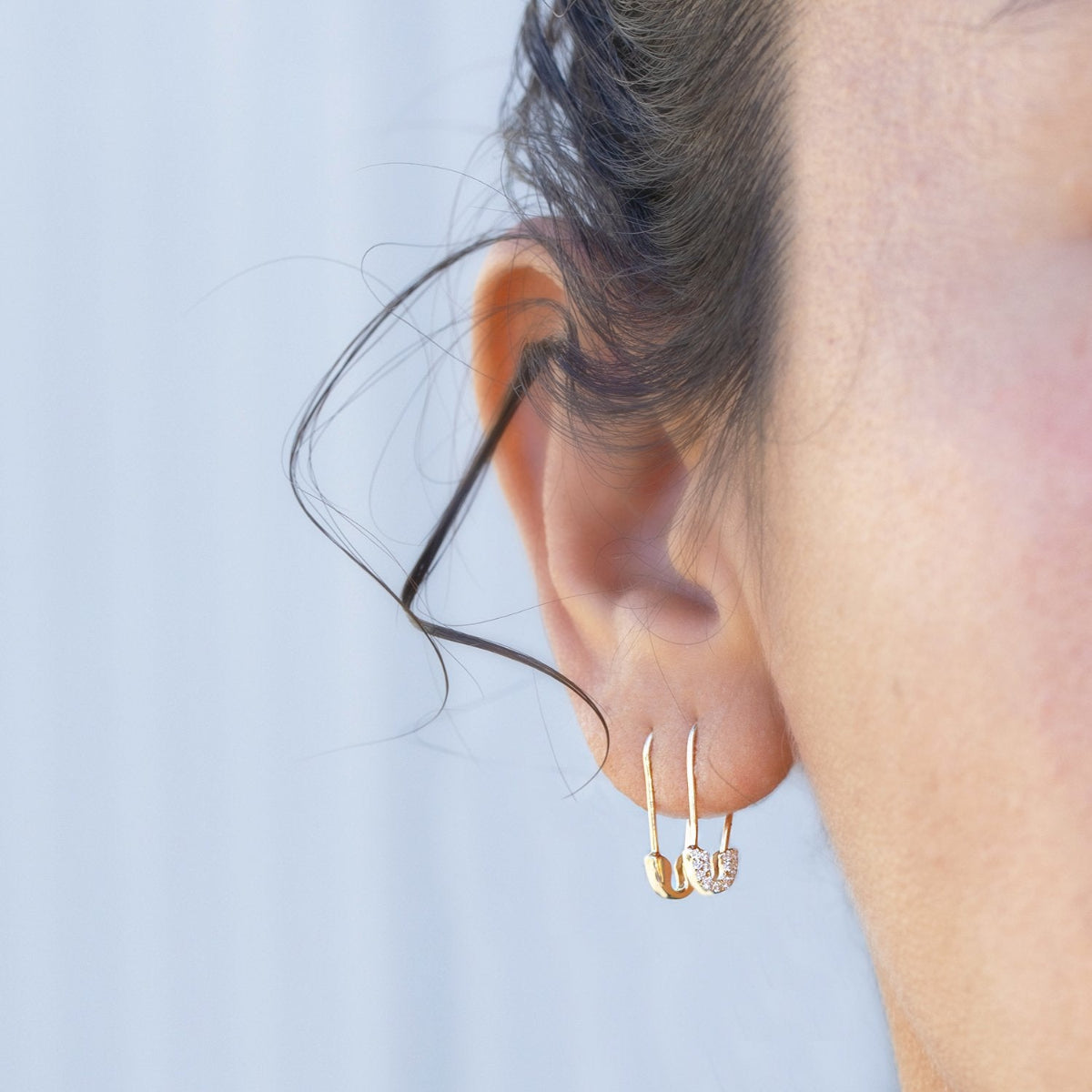 A woman wearing a gold safety pin earring as well as a pave gold safety pin earring on a neutral background