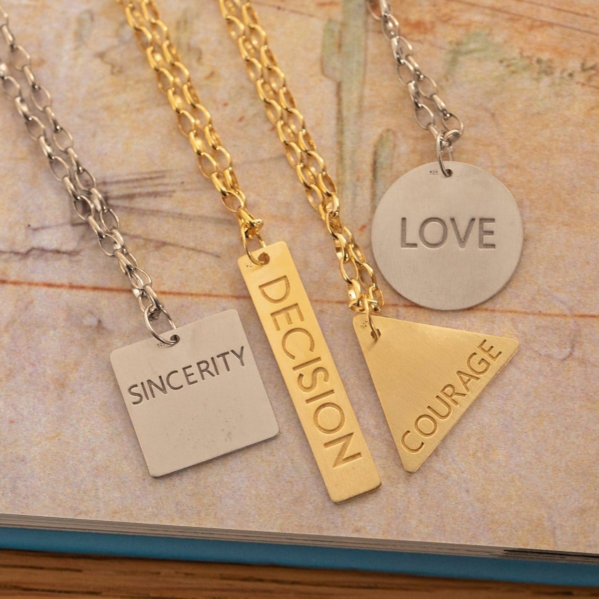 Four Virtues Pendants - Square with the word SINCERITY engraved and Circle with the word LOVE engraved in sterling silver, and Triangle with the word COURAGE and Rectangle with the word DECISION engraved in yellow gold vermeil on top of a page of a book. 