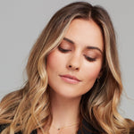 A blonde woman wearing a Single Birthstone Layering Necklace - Yellow Gold in Onyx - August with a Charmed I'm Sure Hoop against a light grey background. 