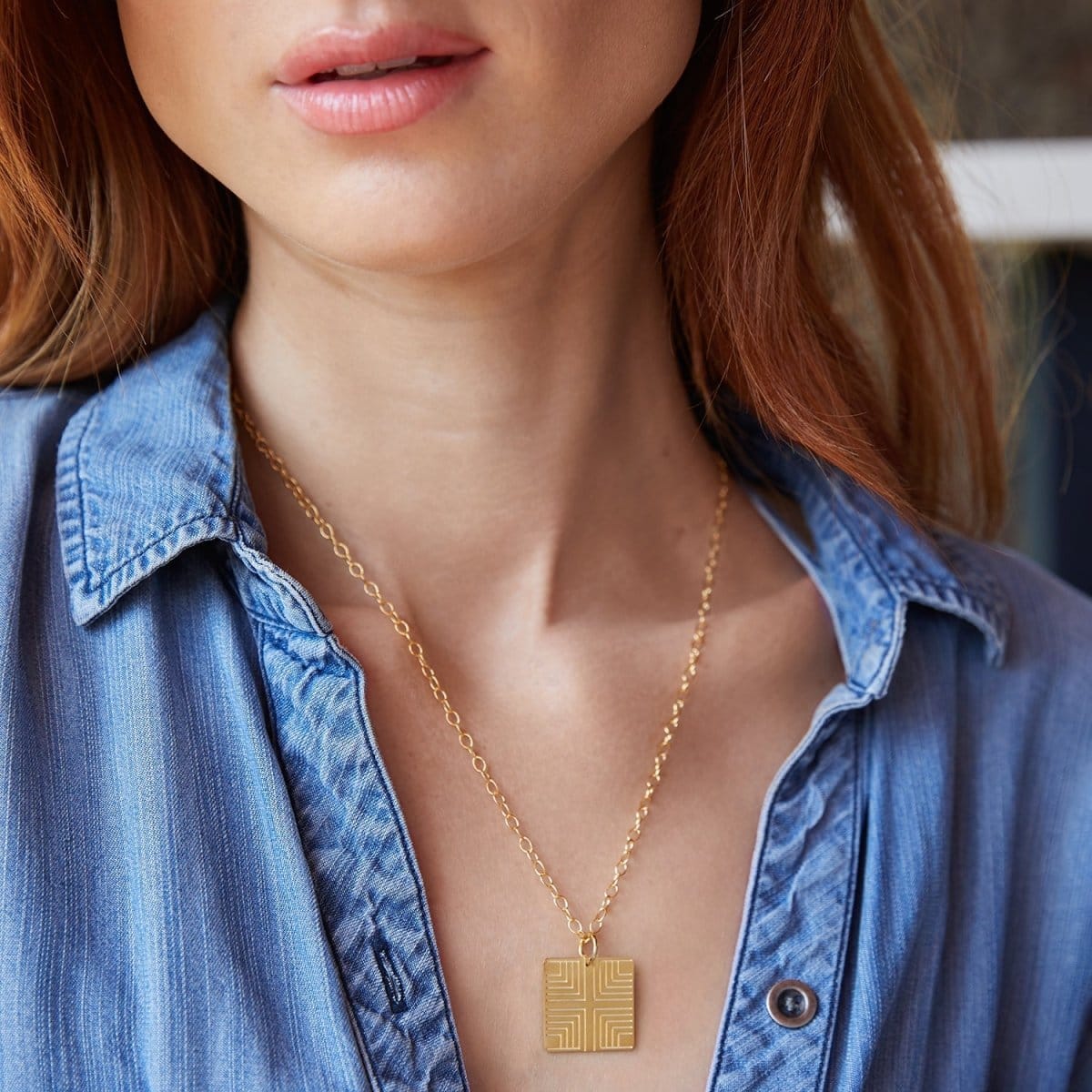 Close-up of a woman's neck wearing a Virtues Pendant - Square in yellow gold with a blue denim shirt against a blurred background. 