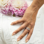 A hand wearing The Forget Me Not Bouquet Ring, The Peony Bouquet Ring and The Magnolia Bouquet Ring in front of a white denim pocket, with pink flowers in the pocket. 