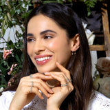 A woman wearing multiple Bouquet Rings, a Gold Safety Pin Earring and Pave Safety Pin Earring, with a white crochet top and brown hair, against a floral background. 