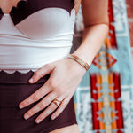 A close-up of a woman’s hand wearing mixed metal Simple Bangles, a rose gold Geo Stacking Ring (Triangle) and a Pavé Geo Ring, with a black and white bodysuit and a colourful blurred background. 