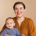 A woman wearing a 14k Gold Asymmetrical Charm Necklace - Star of David and various earrings, styled in a burnt orange top, holding a baby wearing a blue top, against a neutral background. 