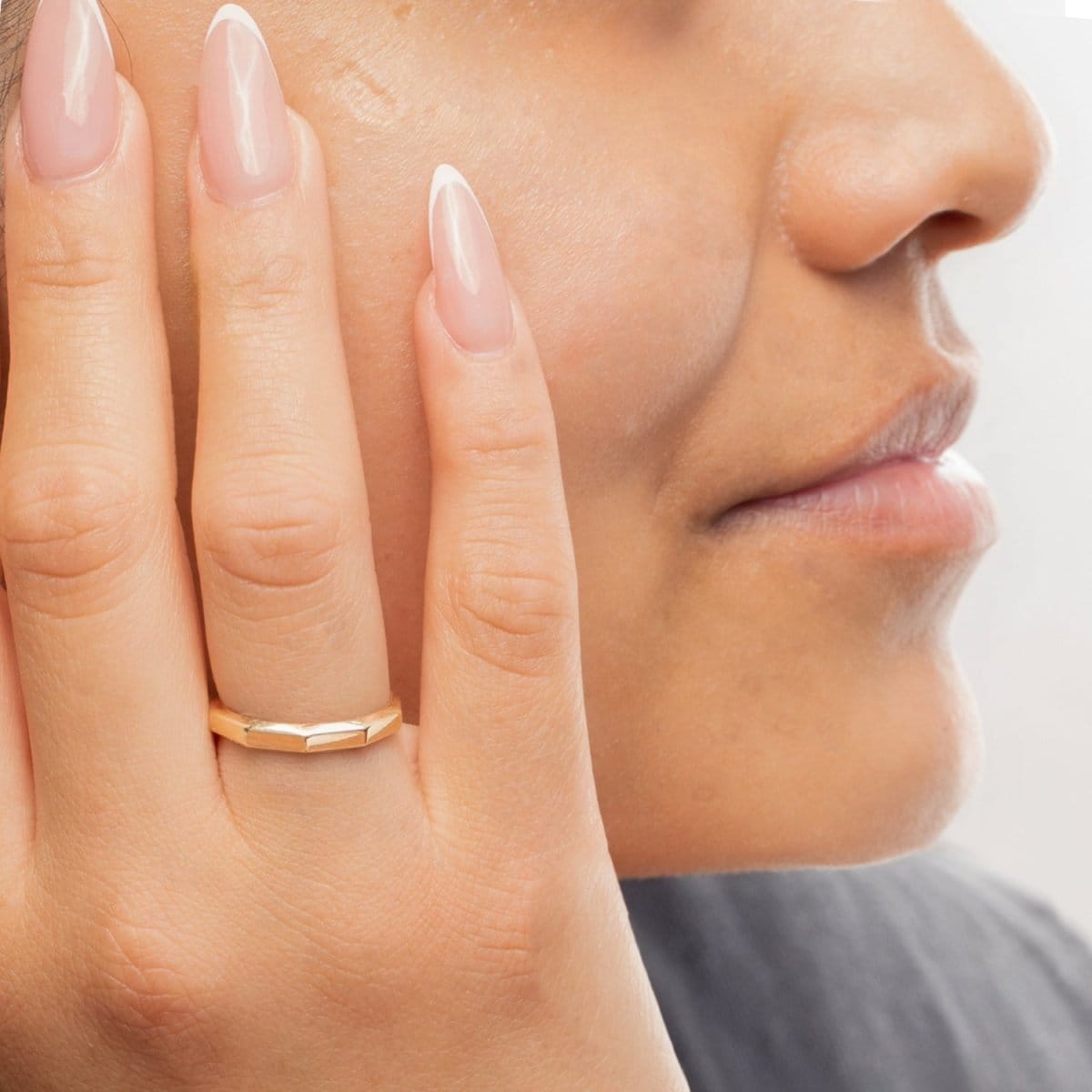 Close-up of a woman's hand wearing a yellow gold Tenfold Ring placed on her face, with a blurred background. 