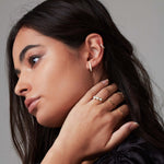 A woman wearing a Tres Pearl Ring, Link Ring, Delicate Twist Band, Flat Ring, Birthstone Ring, Horizon Stud and Pave Ear Cuff with black hair against a light grey background. 