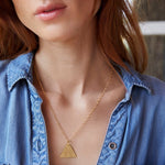 Close-up of a woman's neck wearing a Virtues Pendant - Triangle in yellow gold with a blue denim shirt against a blurred background. 