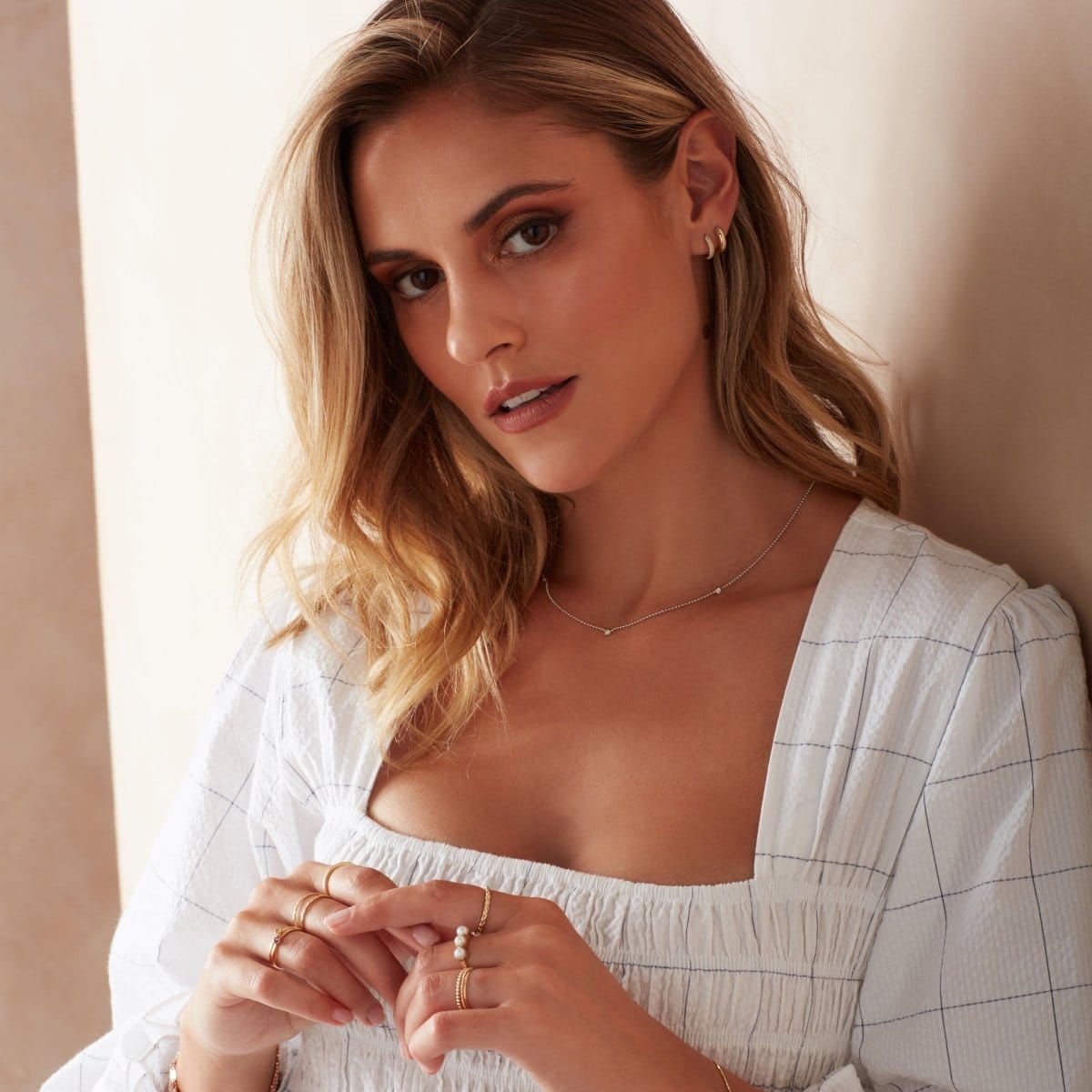 A blonde woman wearing a Triple Birthstone Layering Necklace - White Gold, a Gold Huggie, a Pave Huggie and several rings, including a Tres Pearl Ring, a Braided Ring and Delicate Trio Rings, with a white dress against a neutral background. 