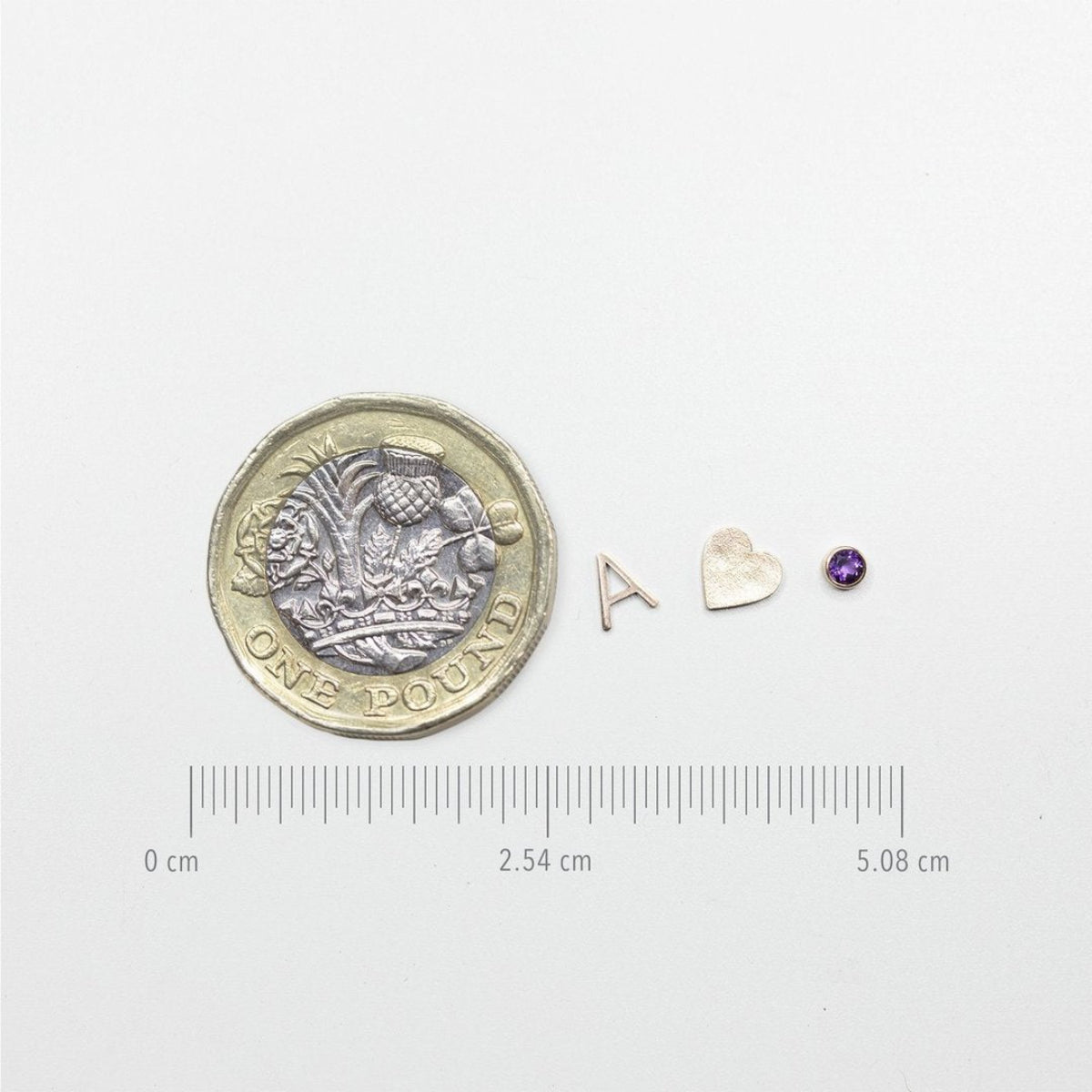 A one pound coin next to an A, heart and birthstone charm with a ruler underneath showing the size of the charms.