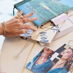 Virtues Pendants - Circle and Square in sterling silver laid out with images and books on a table, alongside a hand wearing a Perla Pinky Ring, Paris Ring, Link Ring and several bracelets, including a Rolo Bracelet and a Love Set Match Bracelet.