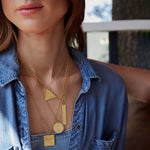 A woman wearing several Virtues Pendants in yellow gold (Triangle, Rectangle, Circle and Square)  with a blue denim shirt against a blurred background. 