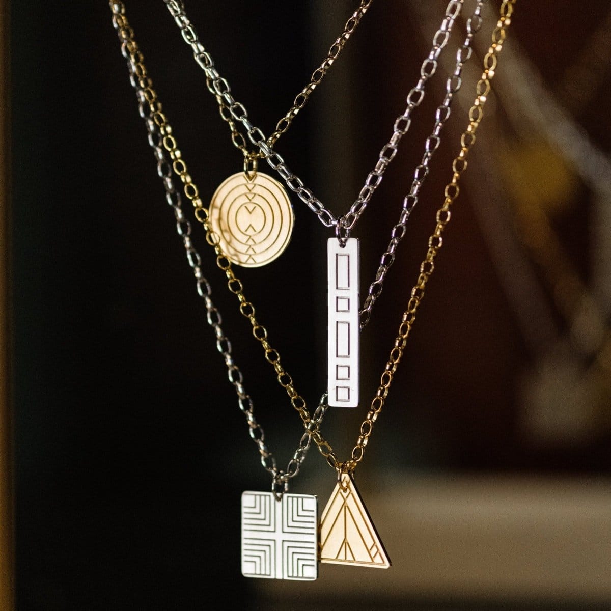 Four Virtues Pendants (Circle and Triangle in yellow gold and Square and Rectangle in sterling silver) hanging against a dark blurred background. 