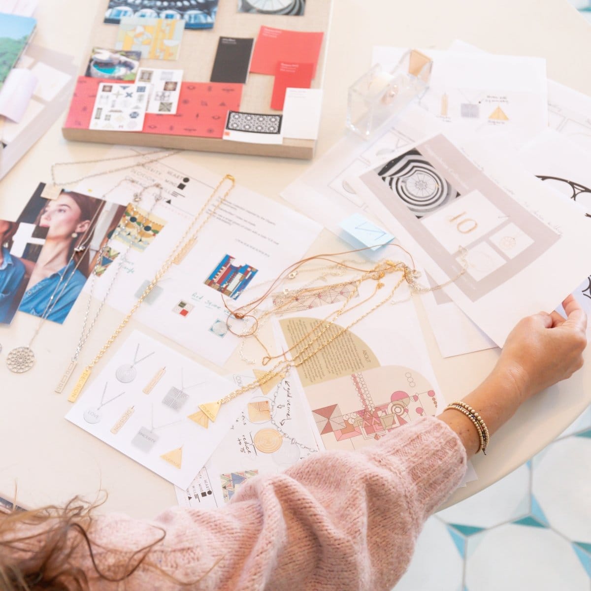 A woman working on jewelry design with Virtues Pendants - Rectangle and Square in yellow gold and sterling silver, a Skylight Necklace in sterling silver laid out with various images. She is wearing Bubble Bracelets and a light pink jumper. 