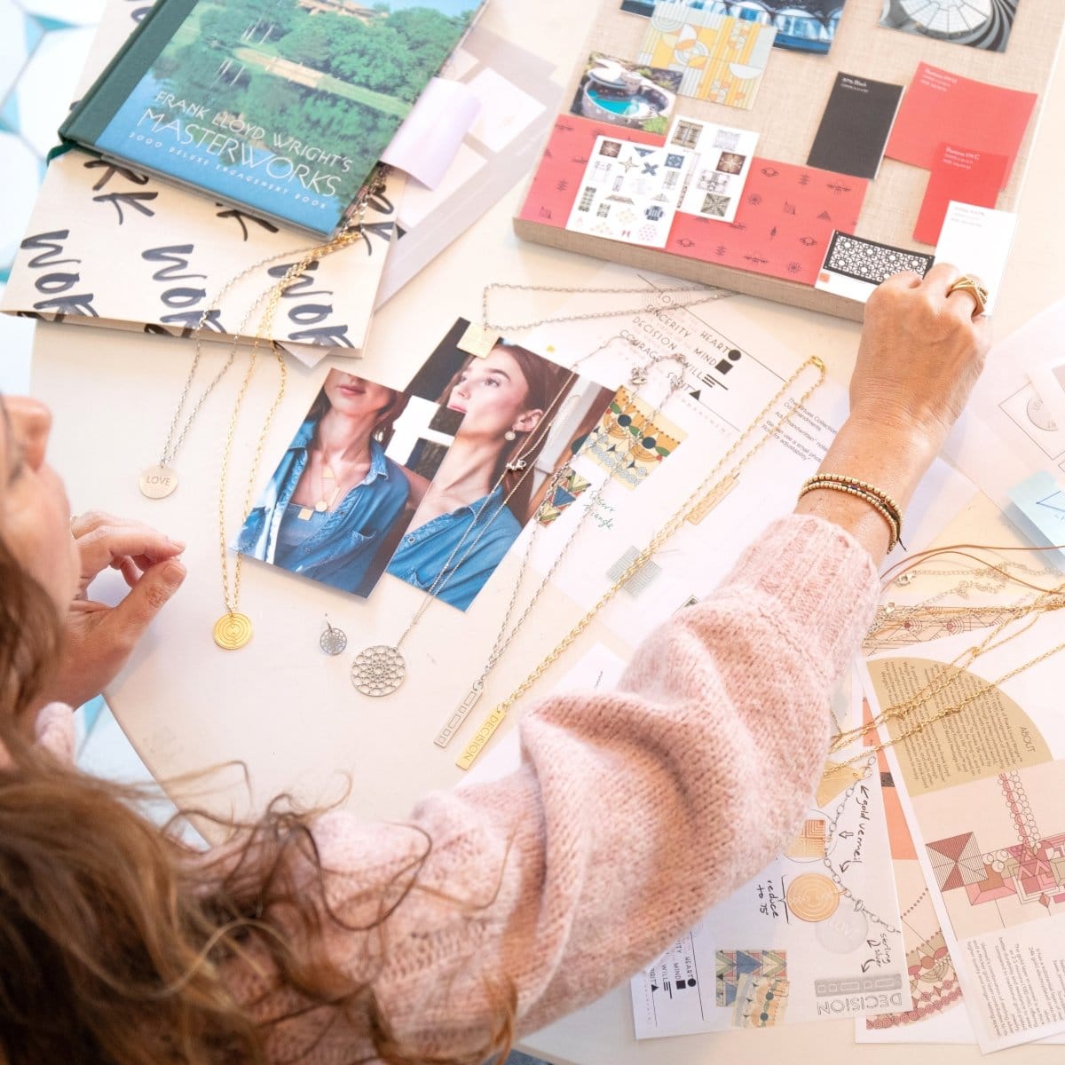 A woman working on jewellery design with Virtues Pendants - Circle and Rectangle in yellow gold and sterling silver, a Skylight Necklace and Hoop in sterling silver. laid out with photos and books. She is wearing Bubble Bracelets. Geo Stacking Rings and a light pink jumper. 