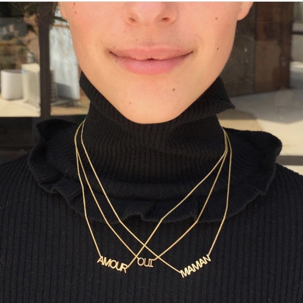 A close-up of a person wearing three yellow gold word plate necklaces in AMOUR, OUI and MAMAN, and a black turtle neck top.