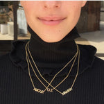A close-up of a person wearing three yellow gold word plate necklaces in AMOUR, OUI and MAMAN, and a black turtle neck top.