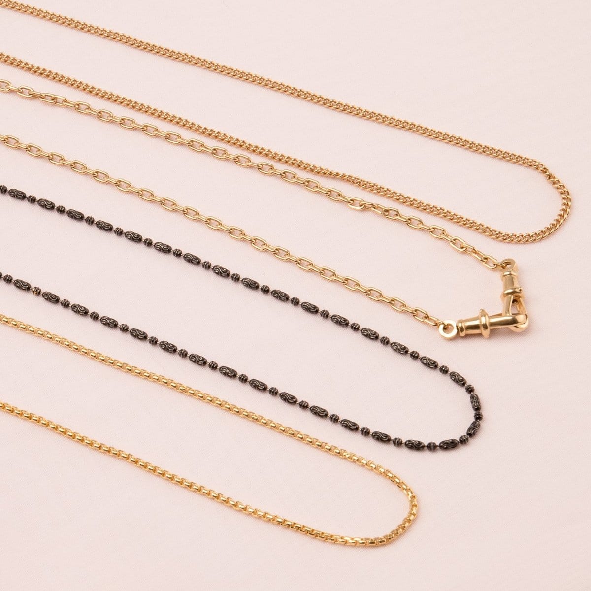 A Black Rhodium Antique Bead Chain, a Round Curb Chain, a Box Chain and a Oval Link Chain laid on an off-white surface. 