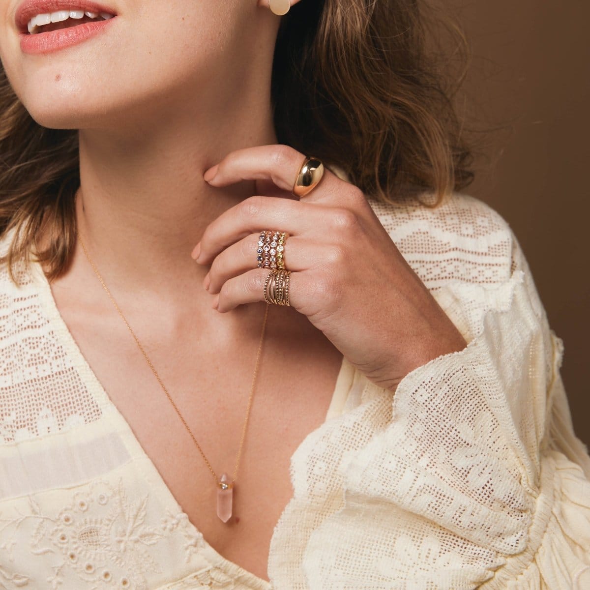 Close-up of a woman wearing a yellow gold Paris Ring, several Bouquet Rings and Delicate Trio Stacking Rings, and a Love Retreat Necklace, styled with a cream lace top against a brown background. 