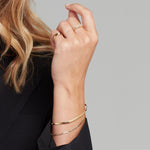 A woman's hand wearing an Overboard Bracelet, another gold bracelet, a Braided Ring, a Delicate Beaded Band and a Perla Pinky Ring, styled with a black blazer. 