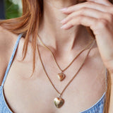 Close-up of a woman's décolletage wearing a Box Chain with a Heart Locket Charm and a Round Curb Chain with a Diamond Heart Locket Charm, all in yellow gold, wearing a denim blue singlet top with her orange hair visible.