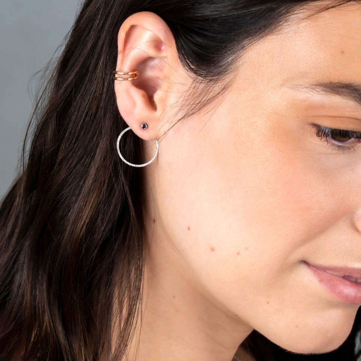 Close-up of a Double Ear Cuff, Birthstone Stud and Circle Ear Jacket Pave in white gold worn on a brunette woman's ear against a light grey background. 