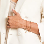 A close-up of a woman’s arm and hand wearing an Off the Cuff Bracelet, a Bubble Bracelet, a Down the Line Bracelet and rings, all in yellow gold, styled with a cream lace top and blazer.