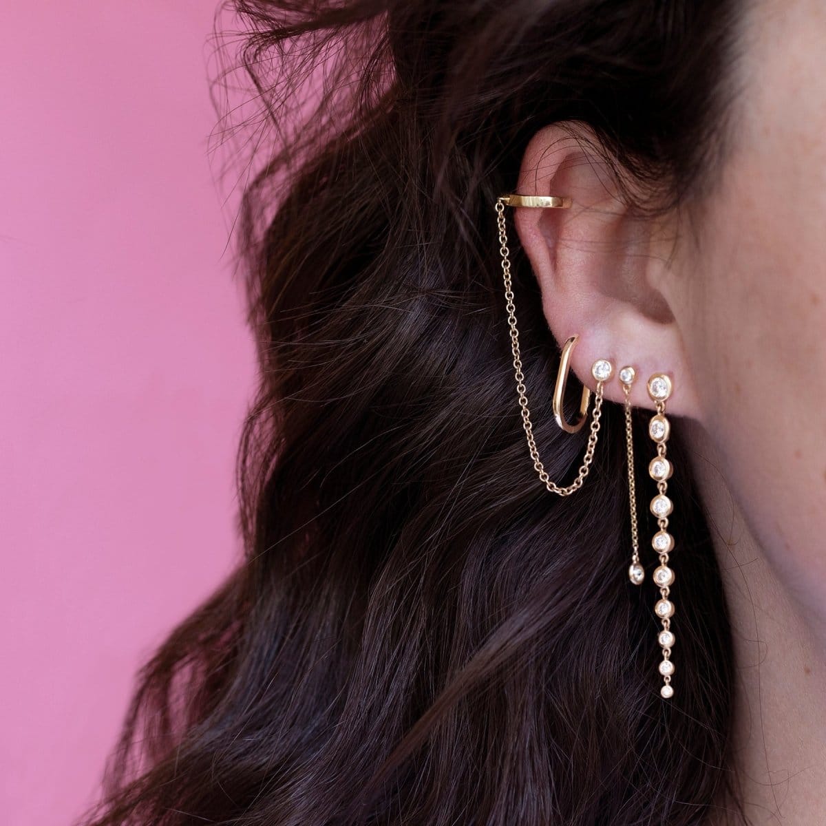 Close-up of an ear with a Diamond Cascade Earring, a Droplet Diamond Earring, a Coupling Ear Cuff + Stud and an Oval Hoop, all in yellow gold, with brown hair and a pink background.