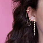 Close-up of an ear with a Diamond Cascade Earring, a Droplet Diamond Earring, a Coupling Ear Cuff + Stud and an Oval Hoop, all in yellow gold, with brown hair and a pink background.