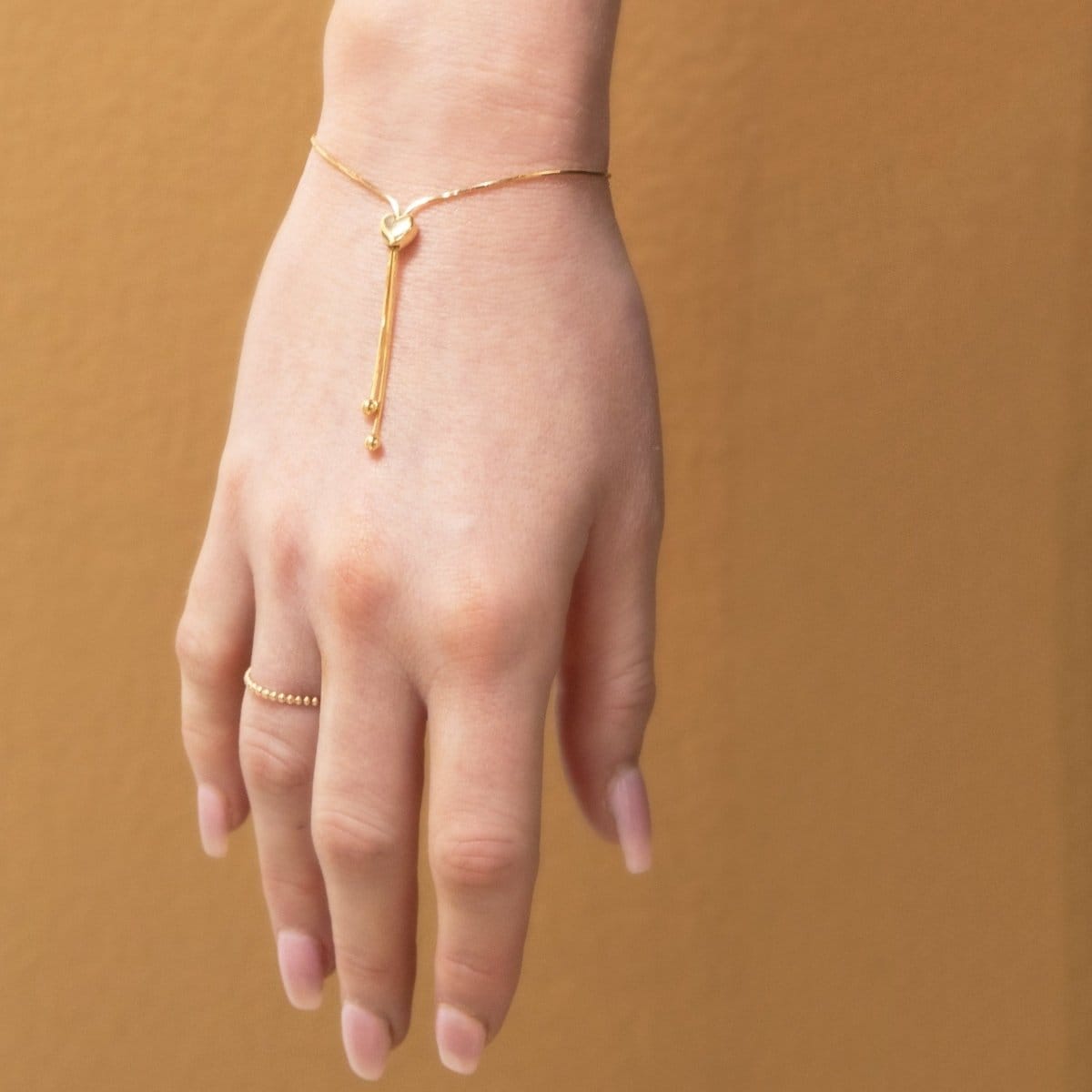 A hand facing down wearing The Slide Heart Bracelet and Delicate Beaded Band in yellow against a tan brown background. 