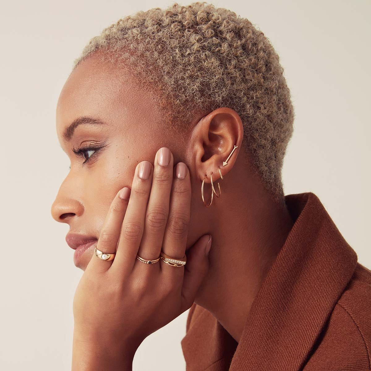 A woman wearing a Crescent Hoop / Medium and Small, Thin Gold Hoop, Straight as an Arrow Earring, Perla Pinky Ring, Birthstone Ring, Delicate Twist Band,  Link Ring and Pave Geo Ring looking to the side with short blonde hair and a brown jacket, against a light neutral background. 