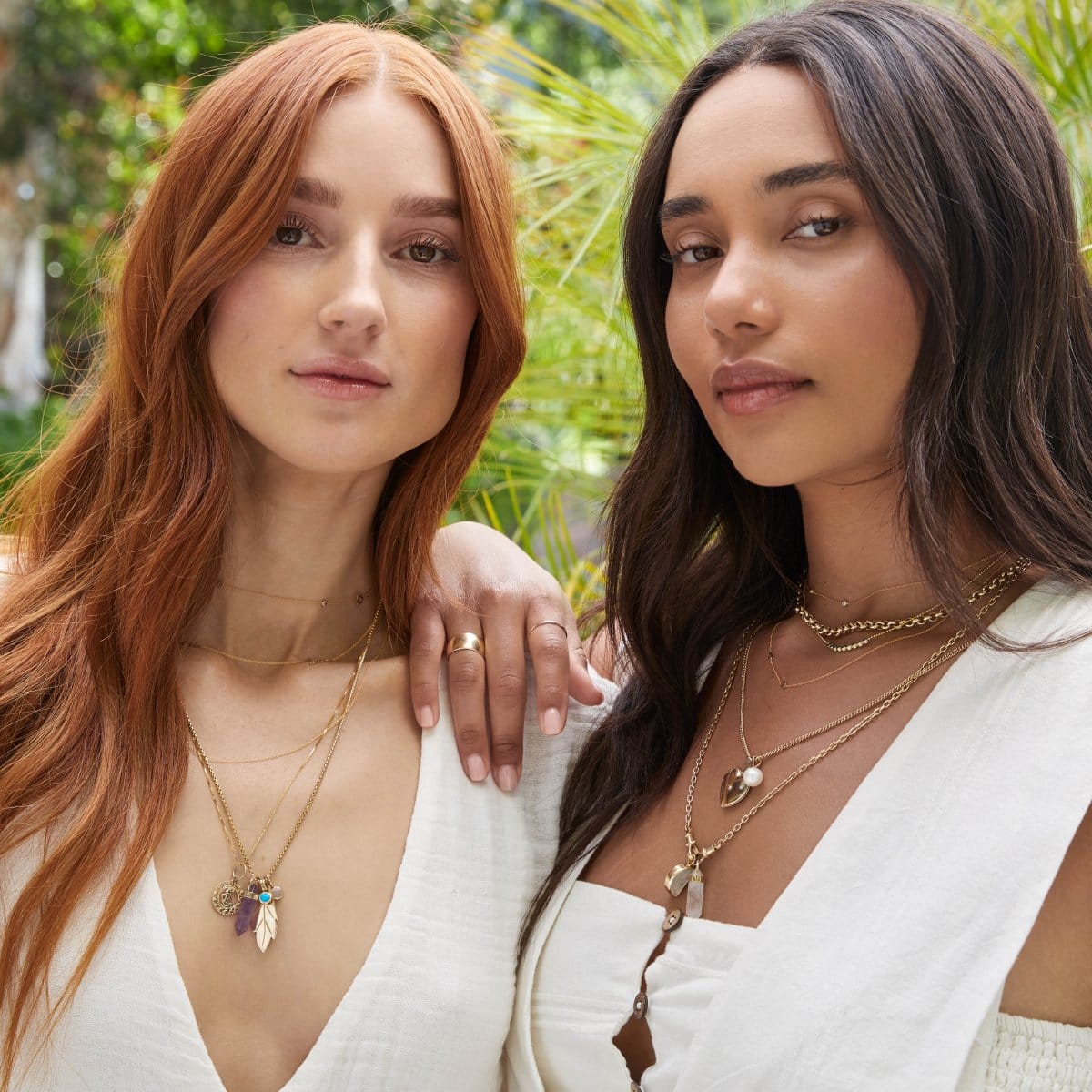 Two women, one wearing Custom Gold Necklaces, The Slide Chain with a Zodiac Pendant, a Box Chain with a Balance Crystal Charm, Turquoise Dangle Charm, Opal Dangle Charm and 20th Anniversary Leaf Pendant, and the other wearing Custom Gold Necklaces, a Rolo Necklace, Diamond Arc Layering Necklace, a Round Curb Chain with a Perla Pendant and Diamond Heart Locket Charm, and an Oval Link Chain with Balance Crystal Charm and Moonstone Crescent Charm, both in white tops, against a blurred leafy background. 