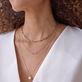 Close-up of a woman's décolletage wearing a yellow gold Bold Link Chain, a Pavé MOM Necklace and a Perla Necklace, with brown curly hair and a white top.