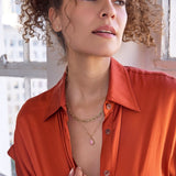 A woman wearing a yellow gold Bold Link Chain, an Element Short Link Chain with a Pink Opal Tear Drop Charm, and a Pavé Charm Stud - Paw, with an orange shirt and brown curly hair against a blurred indoor background.