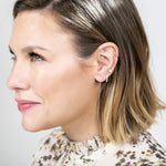 Close-up of an Alli Webb Opal Threader, Opal Huggie and Opal Comfort Stud worn by a woman with short blonde hair and a printed top against a light grey background. 