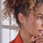 Close-up of a woman wearing a Pavé Charm Stud - Paw and a Link Ring in yellow gold with brown curly hair and a orange top against a light blurred background. 