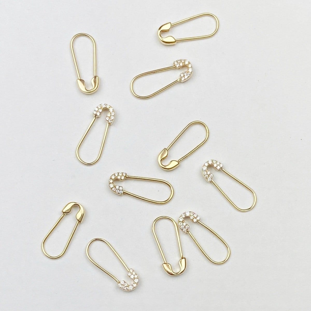 Several Gold Safety Pin Earrings and Pavé Safety Pin Earrings against a light grey background. 