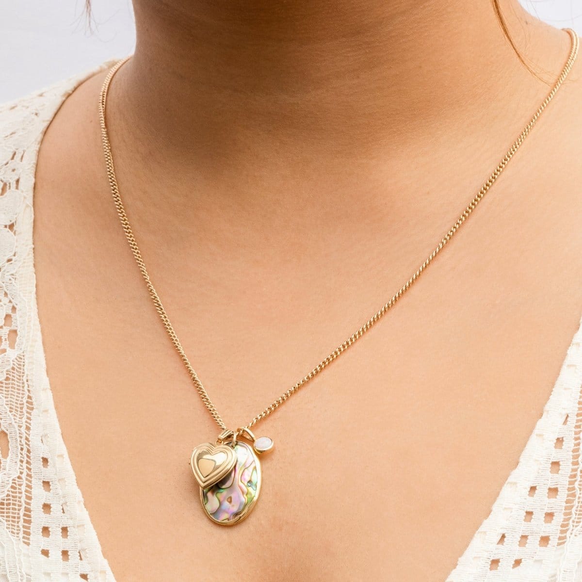 Close-up of a person's décolletage wearing a Round Curb Chain with a Heart Locket Charm, a Large Abalone Cabochon Charm and an Opal Dangle Charm in a cream top.