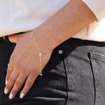 A close-up of a woman’s arm resting on her black jeans, wearing The Slide Heart Bracelet, an Open Diamond Ring and another gold ring, with an off-white top.