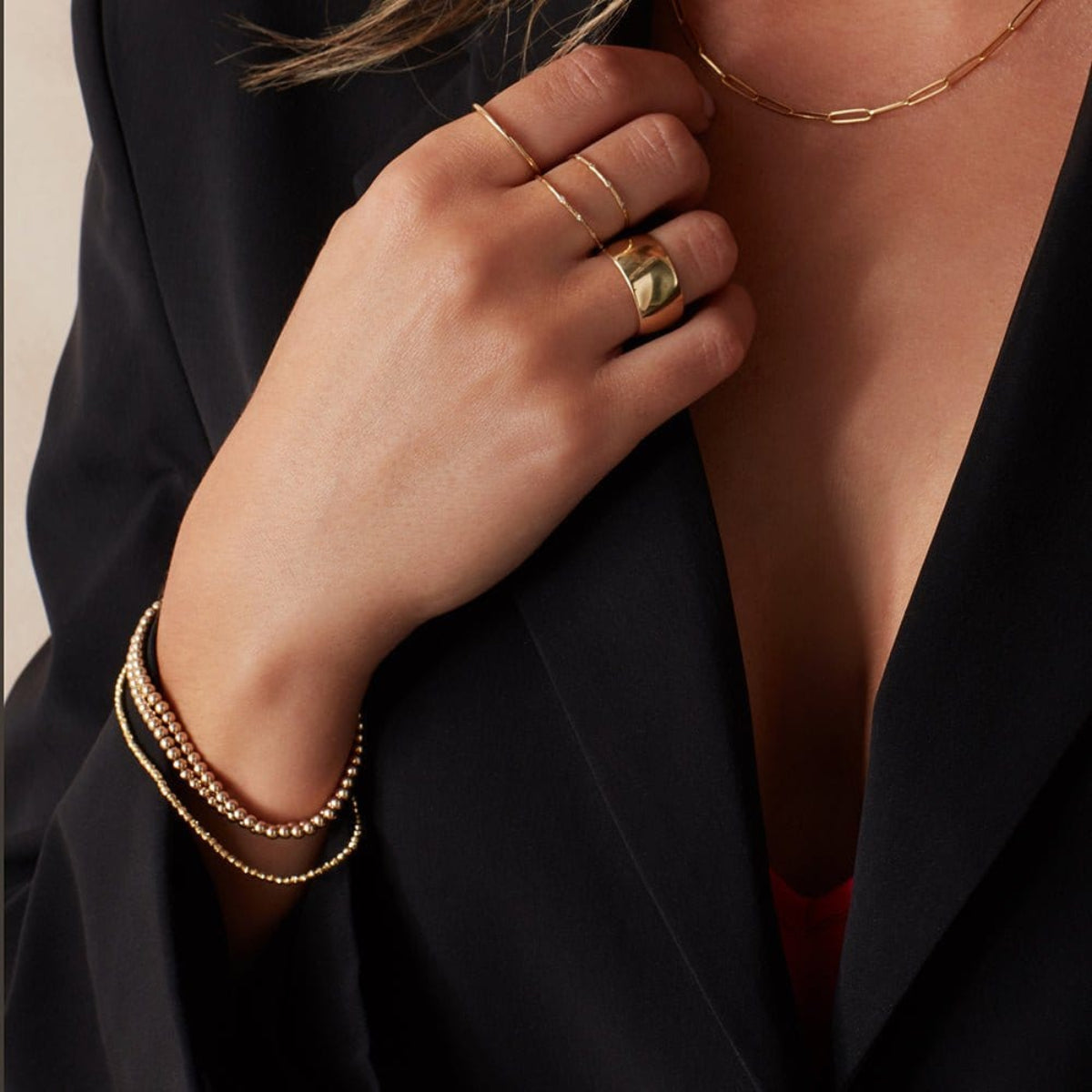 A close-up of a woman’s hand and chest area wearing Bubble Bracelets, a Diamond Cut Bracelet, an Element Long Link Chain, a Delicate Band, two 4 Diamond Rings and a Thick Band, all in yellow gold, styled in a black blazer.