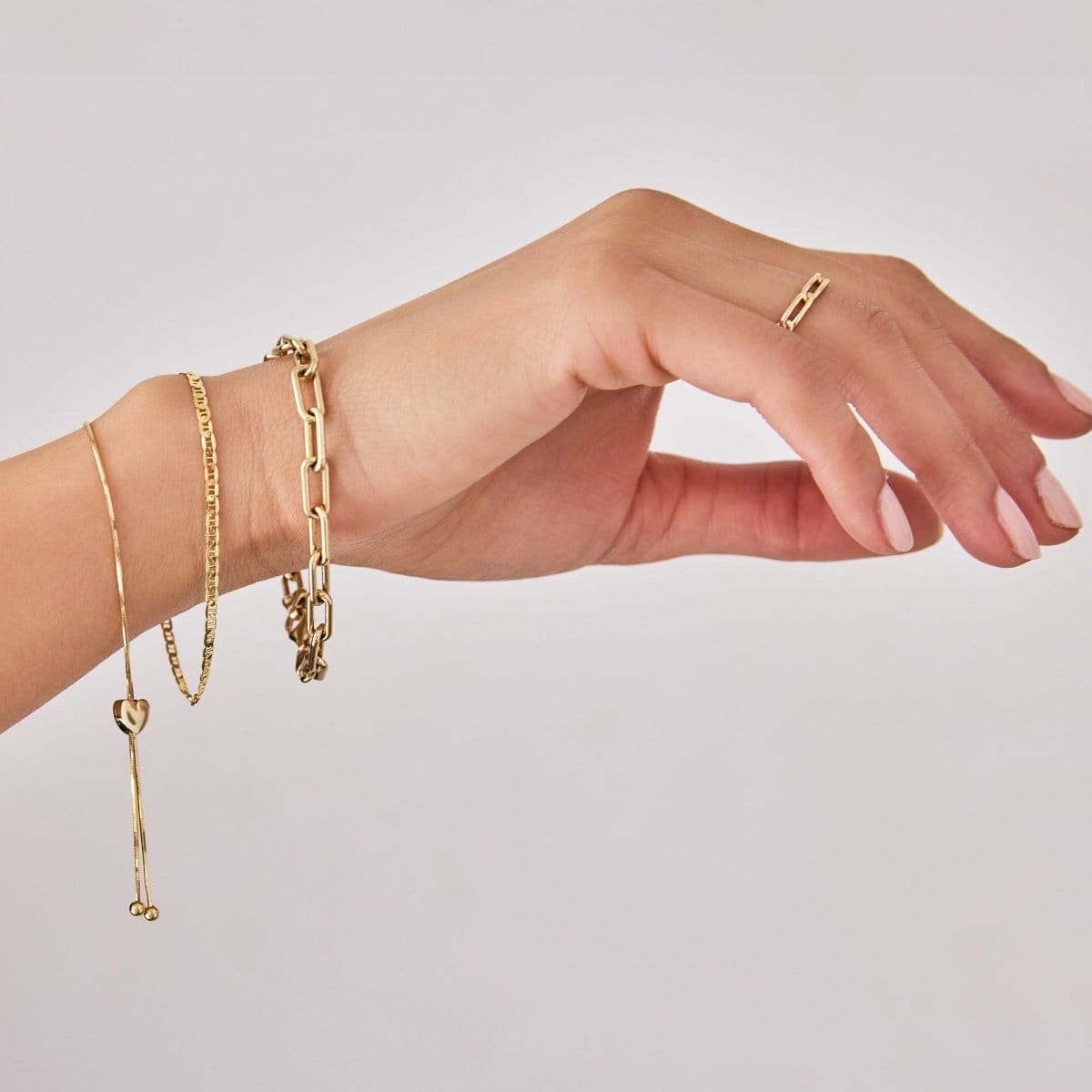 An arm wearing The Slide Heart Bracelet, an Overboard Bracelet, a Bold Link Bracelet and a Link Ring, all in yellow gold, against a light grey background.