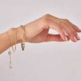 An arm wearing The Slide Heart Bracelet, an Overboard Bracelet, a Bold Link Bracelet and a Link Ring, all in yellow gold, against a light grey background.