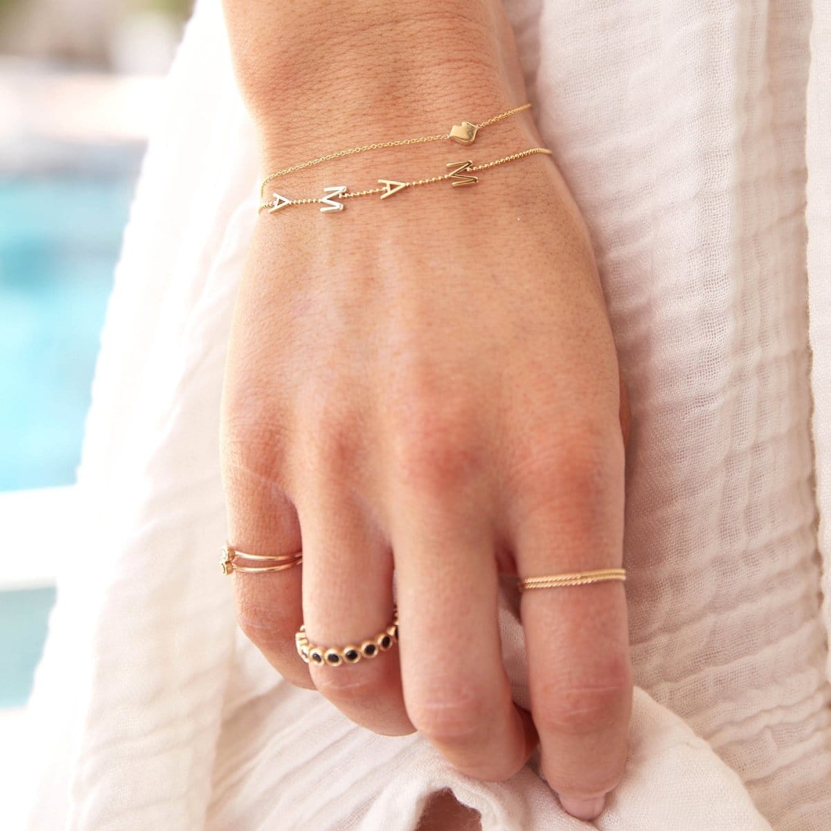 A hand wearing a Lip Bracelet, a Mama Bracelet in a beaded chain, two Birthstone Rings, The Orchard Bouquet Ring and Delicate Twist Bands, all in yellow gold, against a cream fabric background.