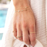 A hand wearing a Lip Bracelet, a Mama Bracelet in a beaded chain, two Birthstone Rings, The Orchard Bouquet Ring and Delicate Twist Bands, all in yellow gold, against a cream fabric background.