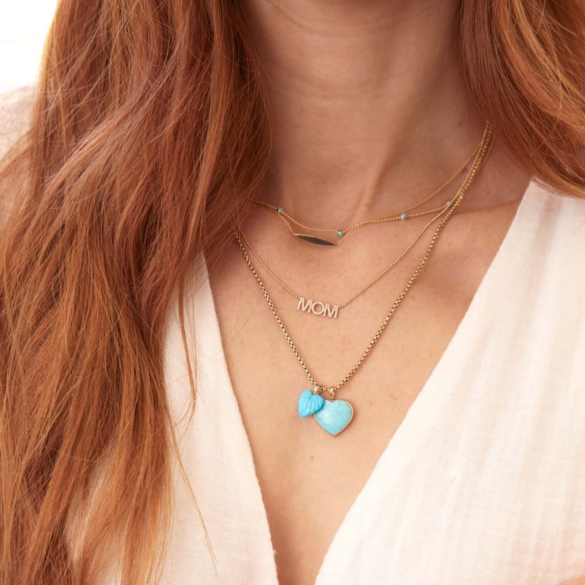 A close-up of a woman's neck wearing a Quintet Birthstone Layering Necklace - Yellow Gold in Turquoise, a Horizon Necklace, a Pavé MUM Necklace, a Box Chain with a Turquoise Heart Charm and an Etched Turquoise Heart Charm, with orange hair styled in a white top.