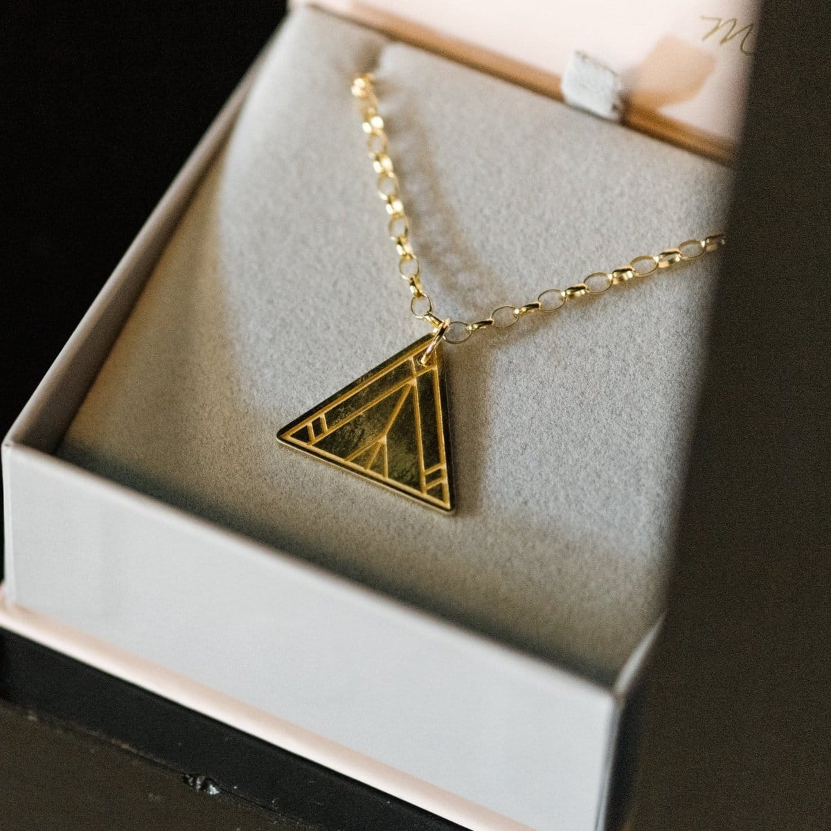 A Virtues Pendant - Triangle in yellow gold in a Maya Brenner light grey and pink jewellery box against a black background. 