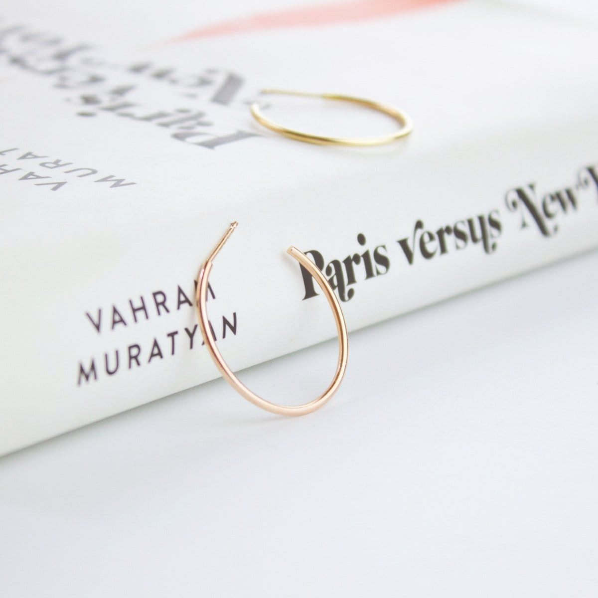 Two Crescent Hoops (one in rose gold and  one in yellow gold) on top of a white book titled Paris versus New York. 
